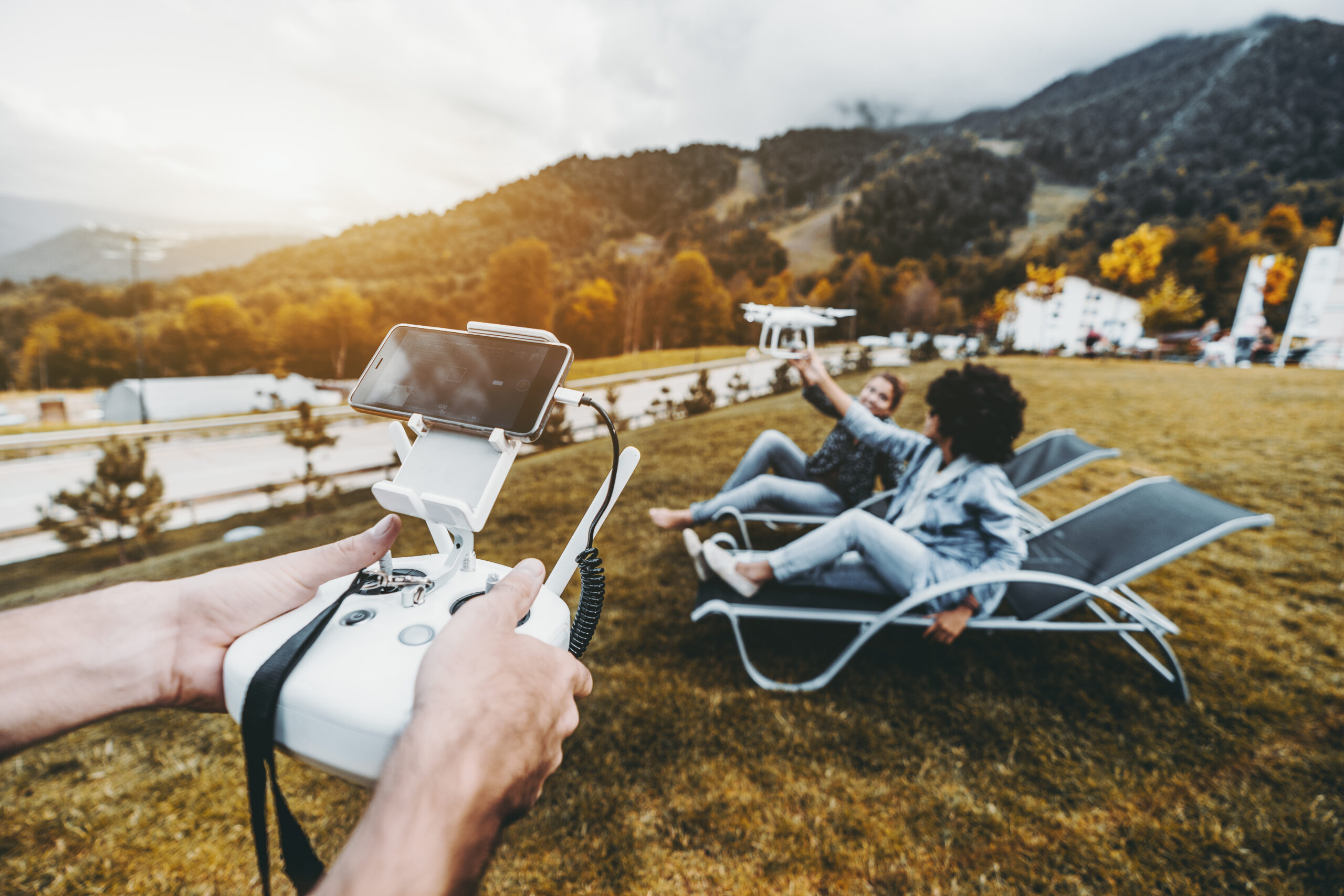 Male hands in foreground holding the remote controller for flying drone operation and two girls in the defocused background laying on deck chairs and touching this multicopter while it filming them